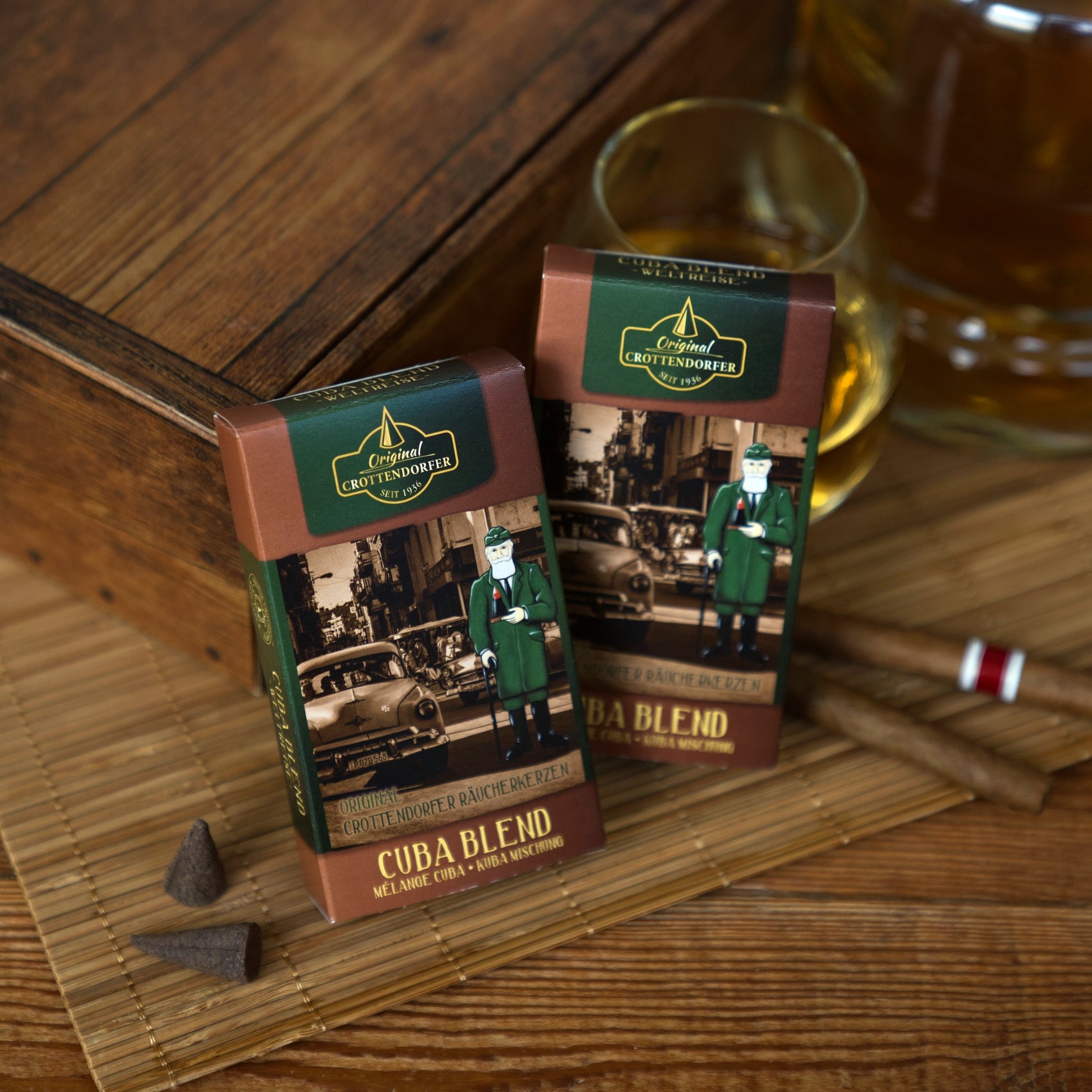 Zwei Schachteln mit Original Crottendorfer Räucherkerzen „Cuba Blend“ im Oldtimer-Design stehen neben einer Holzkiste auf einer Bambusmatte neben einem Glas Whisky, Zigarren und Schokoladenstückchen. Der dezente Räucherduft kubanischer Zigarren und vollmundiger Tabakblätter, den diese Räucherkerzen verströmen, rundet diese elegante Szene ab.