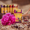 Eine violette „Räucherkerzen Wild Romance“-Box von Original Crottendorfer Räucherkerzen mit Feigen und Granatäpfeln ist umgeben von einer rosa Pfingstrose, Trockenblumen und einem Glasbehälter mit roten Perlen auf einer strukturierten, mediterran anmutenden Oberfläche, mit weiteren verschwommenen Boxen im Hintergrund.