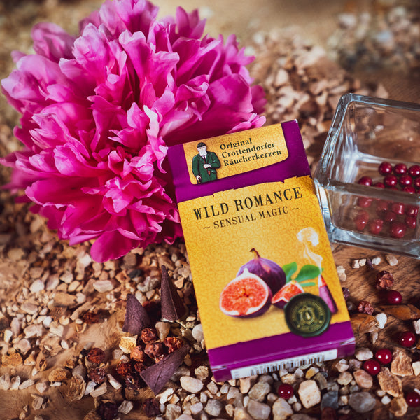 Eine Schachtel „Räucherkerzen Wild Romance“ von Original Crottendorfer Räucherkerzen mit einer Feigenillustration steht auf einer strukturierten Oberfläche mit getrockneten Blumen und Gewürzen. Eine leuchtend rosa Pfingstrose und ein Glasbehälter mit roten Perlen fangen den Charme des nahegelegenen Mittelmeerraums ein.