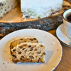 Zwei Scheiben Original Crottendorfer Raacherkerzl-Christstollen mit Rosinen, bestäubt mit Puderzucker, liegen auf einem weißen Teller neben dem Kaffee. Im Hintergrund vervollständigt ein größerer Stollen diese gemütliche Frühstücks- oder Dessertszene.