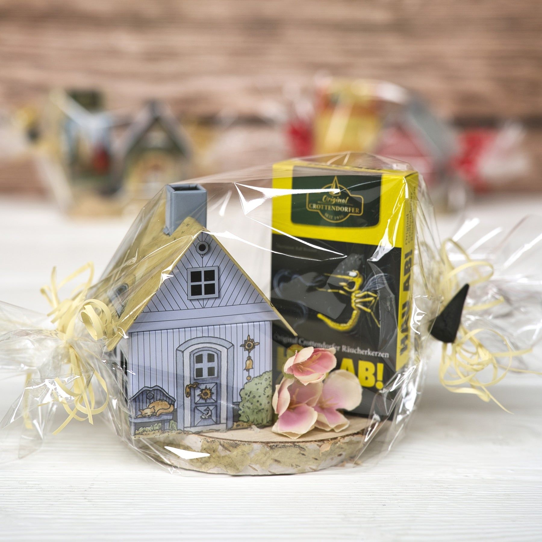 Das Geschenkset „Räucherhäuschen + Räucherkerzen“ von Original Crottendorfer Räucherkerzen enthält ein Crottendorfer Räucherhäuschen, eine Schachtel Räucherkerzen und rosa Blumen, alles in durchsichtige Folie verpackt und auf hellem Holz arrangiert.