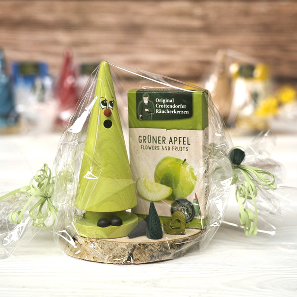 Eine grüne Holzfigur in Form eines Baumes mit Gesicht steht auf einem Holzsockel und ist in durchsichtige Plastikfolie eingewickelt. Daneben steht eine Schachtel mit der Aufschrift „Geschenkset Ziegenbein + Räucherkerzen“ von Original Crottendorfer Räucherkerzen – ein bezauberndes Geschenkset.