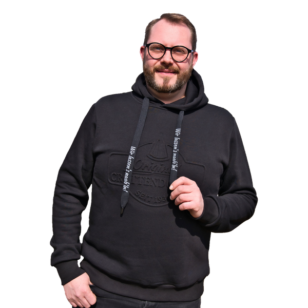 Vor rotem Hintergrund steht ein selbstbewusst lächelnder Mann, der einen Original Crottendorfer Kapuzenpullover mit Crottendorfer Räucherkerzen trägt und durch seine stylische Brille einen exklusiven Streetwear-Look besticht.