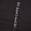 Eine Nahaufnahme des schwarzen strukturierten Stoffes des Original Crottendorfer Kapuzenpullovers zeigt ein geprägtes Logo und einen Riemen mit weißem deutschem Text: „Wir lassens kraabbln!“. Ideal für einen Streetwear-Look, deutet der strapazierfähige Stoff Robustheit an und gibt gleichzeitig teilweise den Blick auf das Logo-Design frei.