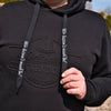 Eine Person trägt einen schwarzen Kapuzenpullover mit dem „Original Crottendorfer“-Logo, der einen Streetwear-Look versprüht. Sie zieht an den Kordeln des Hoodies und trägt den Schriftzug „Wir lassens auch fad!“ vor einem Hintergrund aus verschwommenem Grün und architektonischen Elementen.