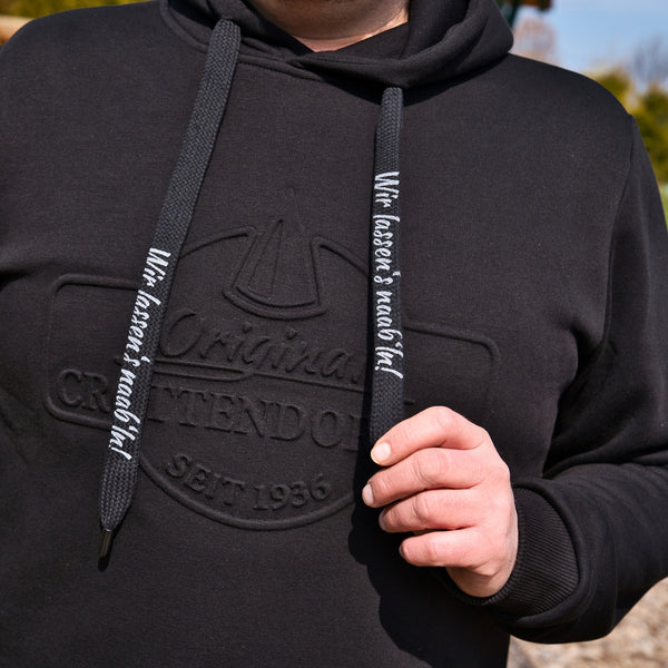 Eine Person trägt einen schwarzen Kapuzenpullover mit dem „Original Crottendorfer“-Logo, der einen Streetwear-Look versprüht. Sie zieht an den Kordeln des Hoodies und trägt den Schriftzug „Wir lassens auch fad!“ vor einem Hintergrund aus verschwommenem Grün und architektonischen Elementen.