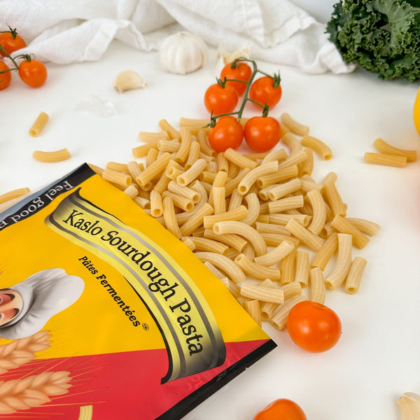 Eine gelbe Miriquidi Sauerteig Pasta-Tüte mit der Aufschrift "Miriquidi Sauerteig Pasta - Makkaroni" verschüttet Makkaroni auf eine weiße Fläche, mit Kirschtomaten, Grünkohl, Knoblauch und einem weißen Tuch im Hintergrund.