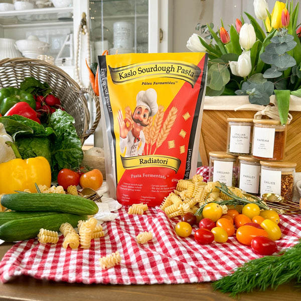 Eine Tüte Miriquidi Sauerteig Pasta - Radiatori steht auf einem Tisch mit frischem Gemüse, Kräutern und Gewürzgläsern. Ein Korb mit Tulpen und ein rot-kariertes Tuch verleihen dieser vegan-freundlichen Pasta-Szene einen dekorativen Touch.