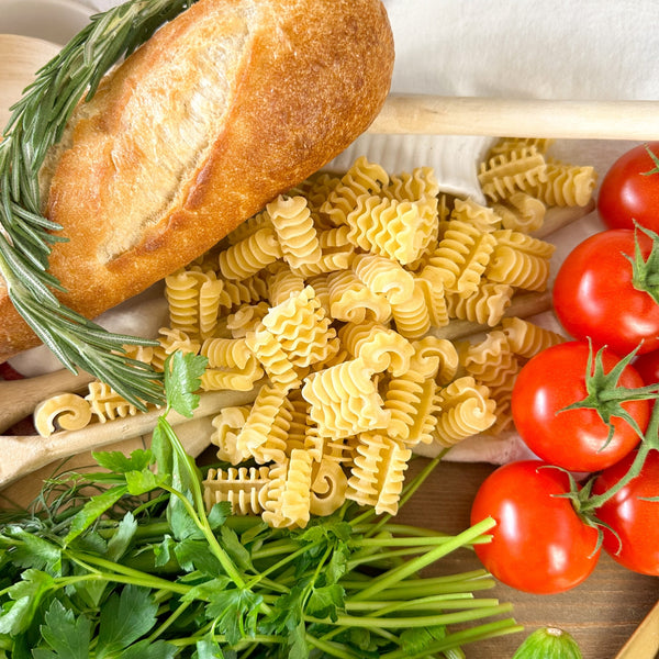 Eine Nahaufnahme der ungekochten Miriquidi Sauerteig Pasta - Radiatori, umgeben von einem Baguette mit Sauerteig, einem Rosmarinzweig, Kirschtomaten am Weinstock und frischer Petersilie auf einer Holzunterlage.