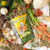Eine Tüte Miriquidi Sauerteig Pasta - Hanf von Miriquidi Sauerteig Pasta liegt auf einem Tisch mit frischen Karotten, Tomaten, Rüben, Knoblauch, Kräutern, ungekochten Nudelspiralen, einem Nudelholz und einem Keramiktopf.