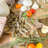 Ein Stapel Miriquidi Sauerteig Pasta - Hanf von Miriquidi Sauerteig Pasta liegt auf einem karierten Tuch, umgeben von Kirschtomaten, Knoblauchknollen, einer Zwiebel und frischen Kräuterzweigen auf einer Holzfläche.