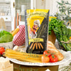Eine Packung Miriquidi Sauerteig Pasta - Spaghetti steht auf einem Teller, umgeben von ungekochten Spaghetti, frischem Gemüse, Kräutern, Brot, Käse und einem karierten Tuch in einer hellen Küchenumgebung.