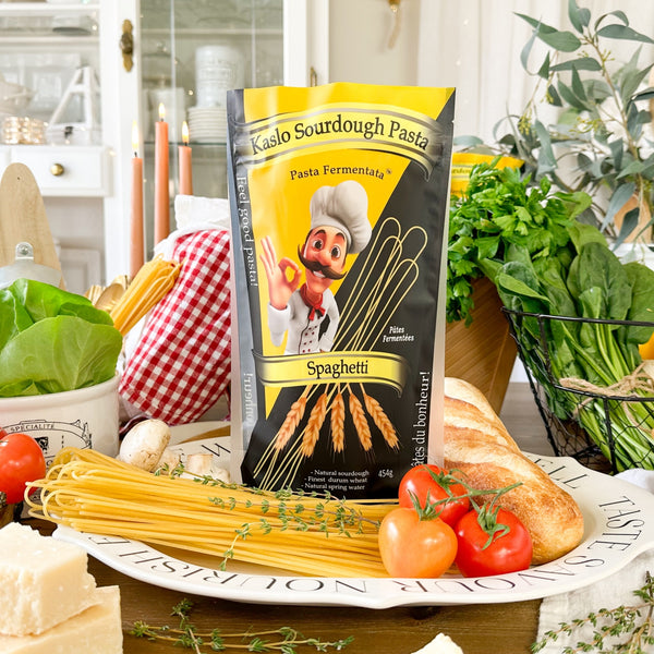 Eine Packung Miriquidi Sauerteig Pasta - Spaghetti steht auf einem Teller, umgeben von ungekochten Spaghetti, frischem Gemüse, Kräutern, Brot, Käse und einem karierten Tuch in einer hellen Küchenumgebung.