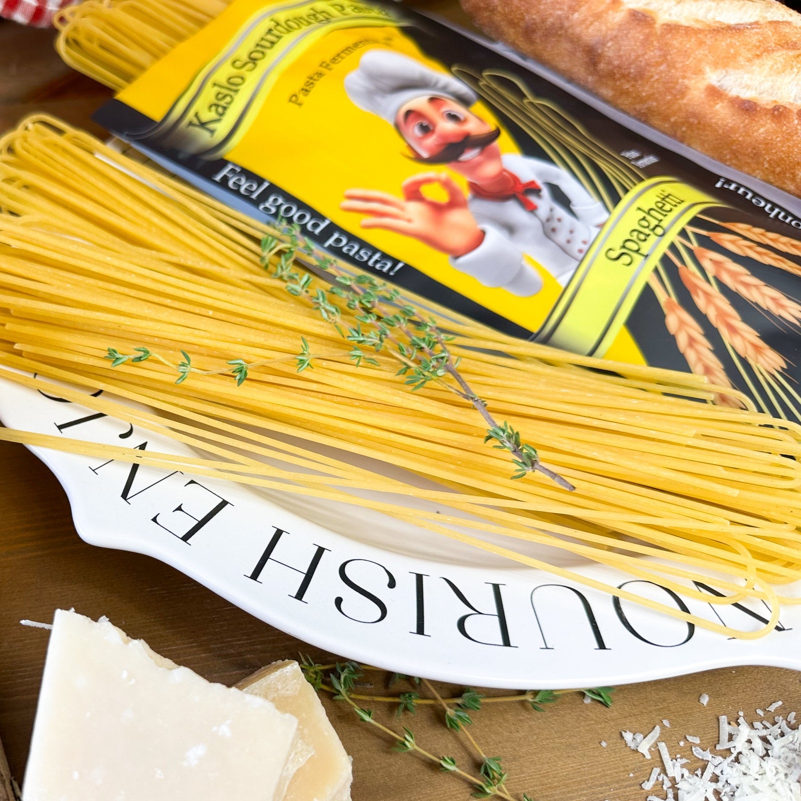 Ungekochte Miriquidi Sauerteig Pasta - Spaghetti auf einem weißen Teller mit der Aufschrift NOURISH ENJ, garniert mit Thymian; daneben eine Nudelpackung mit einem Cartoon-Koch, ein Brotlaib und Parmesankäse. Marke: Miriquidi Sauerteig Nudeln.