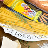 Ungekochte Miriquidi Sauerteig Pasta - Spaghetti auf einem weißen Teller mit der Aufschrift NOURISH ENJ, garniert mit Thymian; daneben eine Nudelpackung mit einem Cartoon-Koch, ein Brotlaib und Parmesankäse. Marke: Miriquidi Sauerteig Nudeln.