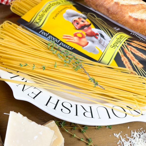 Ungekochte Miriquidi Sauerteig Pasta - Spaghetti auf einem weißen Teller mit der Aufschrift NOURISH ENJ, garniert mit Thymian; daneben eine Nudelpackung mit einem Cartoon-Koch, ein Brotlaib und Parmesankäse. Marke: Miriquidi Sauerteig Nudeln.