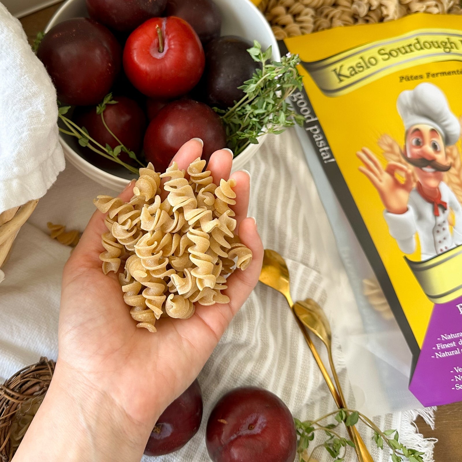Eine Hand hält getrocknete Miriquidi Sauerteig Pasta - Quinoa über eine Schale mit dunkelroten Pflaumen. In der Nähe liegen Thymianzweige, eine gelbe Schachtel Miriquidi Sauerteig Pasta, eine goldene Gabel und weitere Pflaumen auf einem hellen Tischtuch.