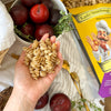 Eine Hand hält getrocknete Miriquidi Sauerteig Pasta - Quinoa über eine Schale mit dunkelroten Pflaumen. In der Nähe liegen Thymianzweige, eine gelbe Schachtel Miriquidi Sauerteig Pasta, eine goldene Gabel und weitere Pflaumen auf einem hellen Tischtuch.