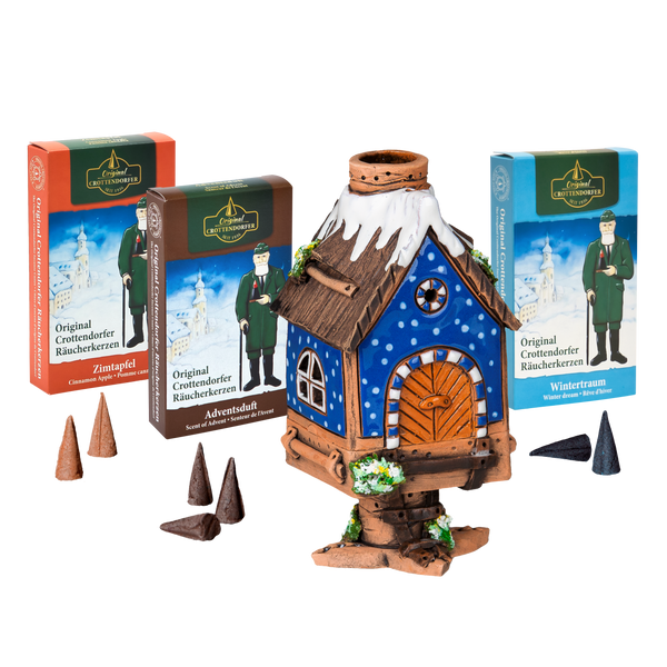 Das Original Crottendorfer Räucherhaus aus Keramik inkl. 3 Packungen Räucherkerzen besticht durch ein blaues Dach mit verschneiten Details und wird mit drei verschiedenen Räucherkerzenpackungen geliefert – ideal für festliche Weihnachtsdekoration vor weißem Hintergrund.