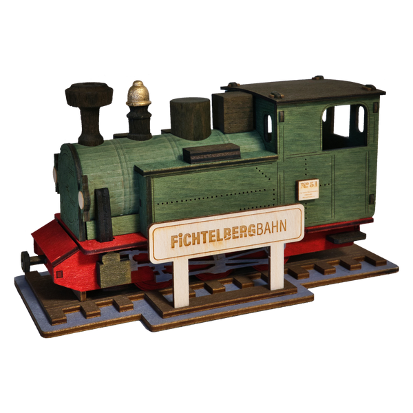 Die Räucherlok Fichtelbergbahn grün von Lenk & Sohn ist eine kleine, grüne Modelleisenbahnlok mit roten Akzenten und einem kastenförmigen, alten Stil - perfekt für Dampflok-Abenteuer. Sie steht auf kurzen Gleisen neben einem Fichtelbergbahn-Schild.