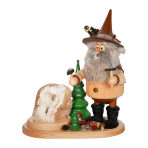 Der Erzwichtel Hauer der DWU Drechselwerkstatt Uhlig ist eine hölzerne Räuchermann-Figur, die einen bärtigen Mann mit spitzem Hut darstellt, der Werkzeuge neben einem grünen Baum und einem Kristallfelsen hält, mit Tannenzapfen und Dekorationen zu seinen Füßen.