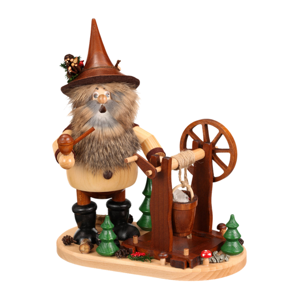 Der Erzwichtel Haspelknecht der DWU Drechselwerkstatt Uhlig ist eine Holzfigur mit braunem Hut, Pfeife und Kunstfellbart, die an einem Brunnen mit Spinnrad inmitten von Mini-Bäumen und Pilzen steht - ein echter erzgebirgischer Räuchermann.