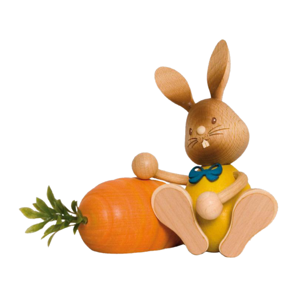Der Stupsi Hase mit Möhre von Drechslerei Kuhnert ist eine hölzerne Hasenfigur in Gelb mit blauer Schleife, die neben einer übergroßen orangefarbenen Karotte mit üppigen grünen Blättern sitzt und aufwendige handgeschnitzte Details und einen fröhlichen Gesichtsausdruck aufweist.