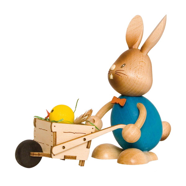 Der Stupsi Hase mit Schubkarre von Drechslerei Kuhnert ist ein bezaubernder Holzhase mit großen Ohren, blauem Oberkörper und orangefarbener Fliege. Er schiebt eine Schubkarre mit einem gelben Küken und Stroh und hat ein schlicht bemaltes Gesicht, das die entzückende Essenz des Osterhasens einfängt.