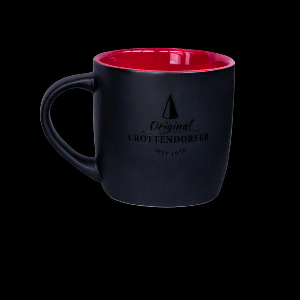 Die Keramiktasse matt rot / schwarz von Original Crottendorfer Räucherkerzen verfügt über eine mattschwarze Außenseite, eine rotbraune Innenseite, den Text „Original Crottendorfer Seit 1946“, ein Dreieckslogo, ist spülmaschinengeeignet und auf einem schwarzen Hintergrund abgebildet.