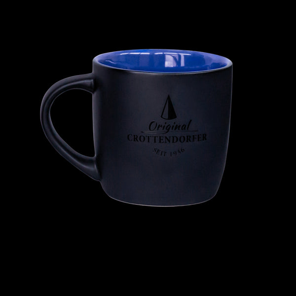 Eine mattblau/schwarze Keramiktasse von Original Crottendorfer Räucherkerzen mit dem Text „Original Crottendorfer Seit 1946“ und einem kleinen Dreieckslogo ist spülmaschinengeeignet und wird vor einem schwarzen Hintergrund angezeigt.