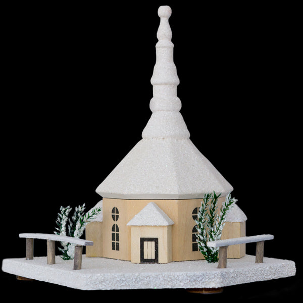 Die Seiffener Kirche, weiß von Original Crottendorfer Räucherkerzen zeigt eine kleine Holzkirche mit einem hohen Kirchturm, grünen Bäumen und Zäunen auf einem schneebedeckten sechseckigen Sockel, die vor einem schwarzen Hintergrund mit weihnachtlichem Charme leuchtet.