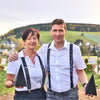 Eine Frau und ein Mann, beide in weißen Hemden und mit dem Aufdruck „Wir lassn’s naab’ln!“ der Original Crottendorfer Hosenträger, stehen nebeneinander im Freien und halten jeweils ein schwarzes Dreieck mit grünen Hügeln und Häusern im Hintergrund.