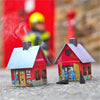 Zwei kleine rote Räucherhäuschen „Feuerwehr“ von Original Crottendorfer Räucherkerzen blasen Rauch auf den Boden. Eine verschwommene Feuerwehrfigur steht neben einer roten Tür und verstärkt den Charme der Szene.