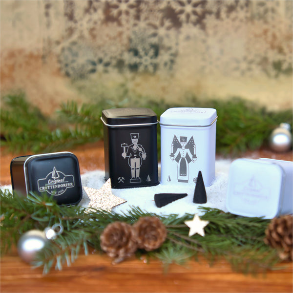 Das quadratische Blechdosen-Set „Bergmann & Engel“ von Original Crottendorfer Räucherkerzen ist kunstvoll mit Tannenzapfen, immergrünen Zweigen und Weihnachtsschmuck auf Schnee arrangiert und schafft so eine zauberhafte Weihnachtszauber-Szene.