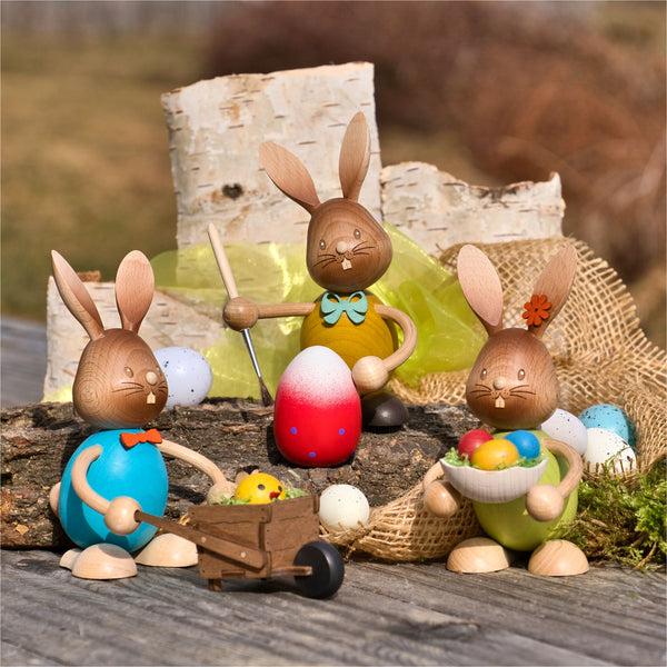Drei aus nachhaltig gewonnenem Holz gefertigte „Stupsi Hase Eiermaler“-Figuren der Drechslerei Kuhnert zeigen einen Hasen, der Eier bemalt, einen anderen, der Eier in einer Schubkarre schiebt, und den dritten, der eine Schüssel mit Eiern zwischen Gras, Moos und einem Birkenstamm auf einer Holzfläche im Freien hält.
